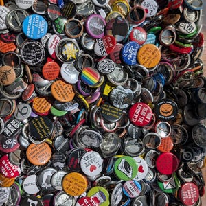 May include: A large collection of circular pin-back buttons in various colors and designs. The buttons feature diverse text phrases such as 'I don't remember your name either', 'Read more zines', 'Don't read the comments', and 'I craft beer'.
