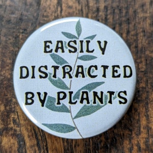 Könnte beinhalten: Ein weißer Knopf mit einem grünen Blatt-Zweig-Design und dem Text "EASILY DISTRACTED BY PLANTS" in schwarzen Buchstaben.