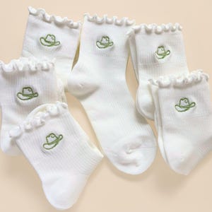 May include: A collection of white ankle socks featuring a ruffled top edge and a small, embroidered green cowboy hat design on the front. The socks are made of a ribbed fabric and are arranged in a group, showcasing the consistent pattern.