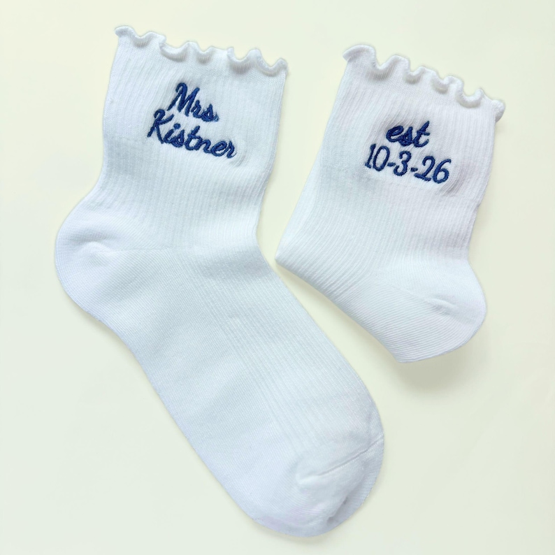 K&ouml;nnte beinhalten: Ein Paar wei&szlig;e Rippsocken mit ger&uuml;schten B&uuml;ndchen. Eine Socke zeigt den dunkelblau gestickten Text Mrs. Kistner, w&auml;hrend die andere Socke den dunkelblau gestickten Text est 10-3-26 aufweist. Die Socken bestehen aus einem weichen Stoff.