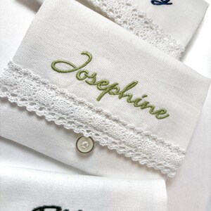 K&ouml;nnte beinhalten: Wei&szlig;e Taschent&uuml;cher mit gestickten Namen und Spitzenbesatz. Auf einem Taschentuch steht der Name "Josephine" in gr&uuml;ner Schrift. Ein kleiner, runder Knopf ist an der Ecke des Taschentuchs befestigt. Weitere Taschent&uuml;cher sind im Hintergrund zu sehen.