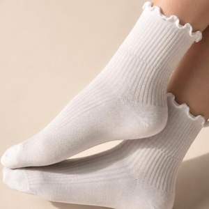 K&ouml;nnte beinhalten: Ein Paar wei&szlig;e Socken mit einer gerippten Strickstruktur am Fu&szlig; und Kn&ouml;chel. Der obere Rand jeder Socke ist mit einem zarten, ger&uuml;schten Saum versehen, der f&uuml;r eine weiche, wellenf&ouml;rmige Optik am oberen B&uuml;ndchen sorgt.