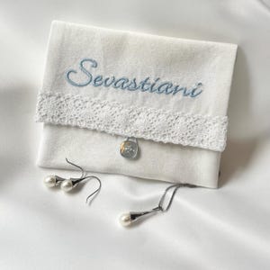 K&ouml;nnte beinhalten: Wei&szlig;e Schmucktasche aus Stoff mit dem Namen "Sevastian" in Hellblau gestickt. Die Tasche hat eine Spitzenborte und einen silbernen Verschluss. Perlenohrringe und eine Perlenkette liegen daneben.