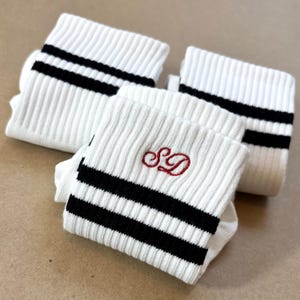 Pode incluir: Meias brancas caneladas com riscas pretas e um monograma "SD" vermelho bordado. As meias est&atilde;o dobradas e empilhadas, mostrando o design. Feitas de um material macio e canelado.