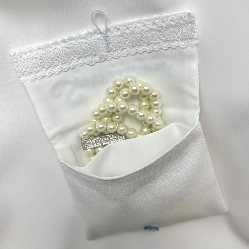 K&ouml;nnte beinhalten: Ein wei&szlig;es Stofft&auml;schchen mit Spitzenbesatz, das ein Perlenarmband mit einem silbernen Verschluss enth&auml;lt. Das T&auml;schchen wird mit einer kleinen Schlaufe und einem Knopf verschlossen. Die Perlen sind cremefarben.