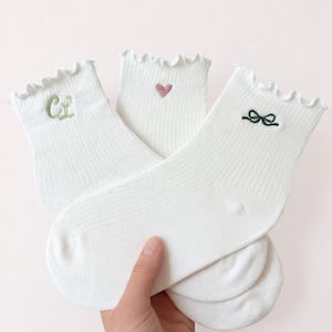 May include: Three pairs of white ankle socks with ruffled edges. One sock has the embroidered letters "CL" in green, another has a pink heart, and the third has a black bow. The socks are made of a ribbed knit fabric.