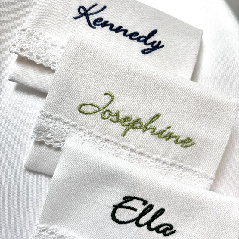 K&ouml;nnte beinhalten: Drei wei&szlig;e Taschent&uuml;cher mit gestickten Namen: "Kennedy" in Marineblau, "Josephine" in Gr&uuml;n und "Ella" in Schwarz. Jedes Taschentuch hat eine zarte Spitzenborte an einer Kante, die einen Hauch von Eleganz verleiht.