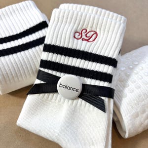 May include: White athletic socks with black stripes and a black ribbon. The socks have the embroidered initials "SD" in red and a white button that says "balance."
