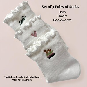May include: A set of three pairs of white socks with frilled edges. Each pair has embroidered designs: a bow, a heart, and a stack of books. The image text reads "Set of 3 Pairs of Socks Bow Heart Bookworm."