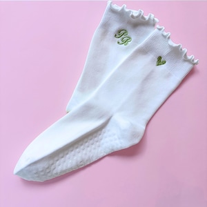 May include: A pair of white ankle socks featuring a ruffled top edge. One sock displays green embroidered letters PR, while the other features a small green embroidered heart. The soles of the socks have a textured grip pattern for traction.