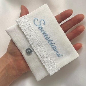 K&ouml;nnte beinhalten: Eine wei&szlig;e Leinentasche mit dem hellblau gestickten Namen "Sebastiani". Die Tasche hat eine zarte Spitzenborte und einen kleinen, dekorativen Knopfverschluss. Die Tasche wird in einer Hand gehalten.