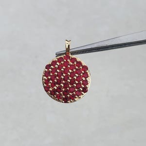 May include: A gold-colored pendant with a circular design, densely covered with small, round, red gemstones. The pendant is held by tweezers against a light gray background. The pendant has a small loop at the top for a chain.