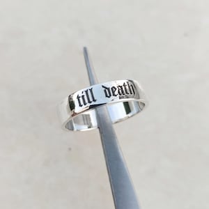 May include: A silver ring with the words "till death" engraved in a gothic font. The ring is held by a silver tool, and the background is a light, neutral color. The ring is a simple band style.
