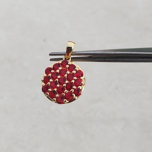 May include: A gold pendant with a circular design, densely set with small, round, red gemstones. The pendant is held by tweezers against a neutral background, showcasing the jewelry's details and craftsmanship.