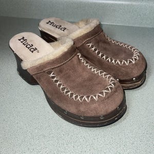 May include: Brown suede clogs with a faux fur lining and white stitching detail. The clogs have a wooden platform sole and the brand name "Mudd" is printed on the insole.