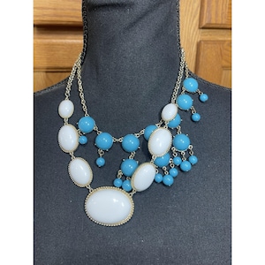 May include: A multi-strand statement necklace featuring a combination of smooth, opaque white oval cabochons and round turquoise blue beads. The design incorporates a gold-tone metal chain link structure that supports the clusters of beads.