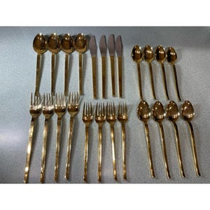May include: A collection of gold-colored flatware, including forks, spoons, and knives. The utensils are arranged on a light-colored surface. The forks have four tines, and the spoons have oval bowls. The knives have a simple, elongated design.