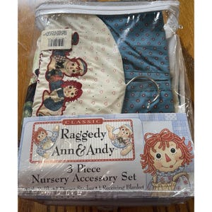 May include: A new, sealed 3-piece nursery accessory set featuring Raggedy Ann & Andy. The set includes a diaper stacker and receiving blanket. The packaging shows illustrations of the characters and the text "Classic Raggedy Ann & Andy".