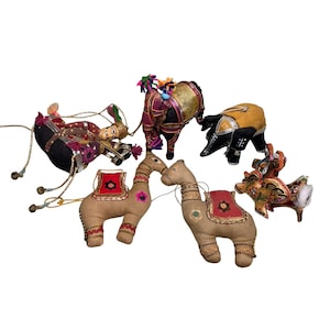 May include: A collection of colorful, handcrafted fabric animal figurines, including camels, a bull, and an elephant. The camels have red and gold saddles. The elephant and bull are adorned with gold and black accents. The figurines are set against a white background.