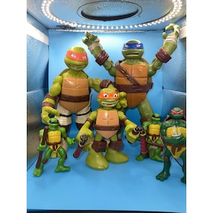 May include: A collection of Teenage Mutant Ninja Turtles action figures. The figures are green with brown shells, each wearing a different colored mask. Some figures are holding weapons. The background is a light blue.