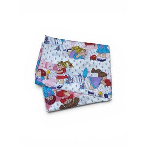 May include: A folded fabric sheet featuring a repeating pattern of cartoon dolls with yarn hair in various outfits, including red dresses and blue tops, set against a white background with small pink flowers, white picket fences, and houses.