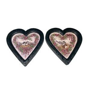 May include: Two heart-shaped wall plaques with black frames. Each plaque features a bird scene with a nest, birds, and floral accents in pink, green, and white. The plaques are displayed against a white background.