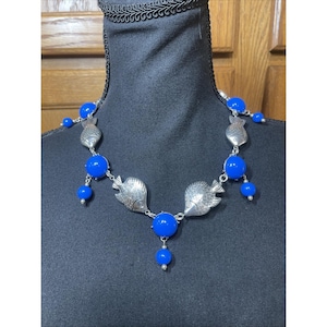 May include: A necklace featuring alternating silver-toned metal fish charms and bright blue spherical beads. The design includes several dangling blue beads suspended from the fish and circular bead links, creating a symmetrical arrangement.