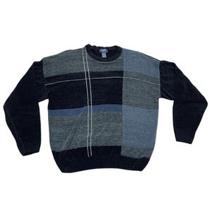 May include: A vintage sweater with a geometric color block design. The sweater features a crew neck and long sleeves. The color scheme includes shades of gray, blue, and black, with white vertical lines. The sweater is made of a soft, textured material.