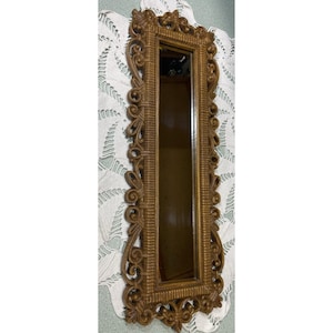 May include: A rectangular wall mirror featuring a brown frame with intricate, raised scrollwork and carved detailing along the edges. The central glass panel is vertically oriented, surrounded by a textured border that mimics woven patterns.