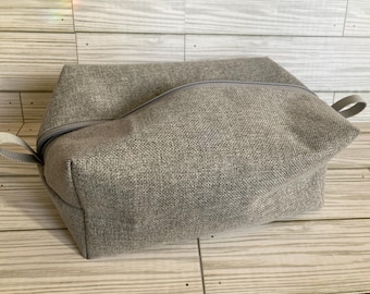 May include: A rectangular gray fabric bag with a curved zipper closure and a small loop handle on one side. The bag is made of a textured material and sits on a wooden surface.