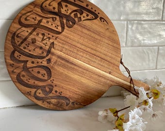 May include: A round wooden serving board with a brown wood grain and a handle. The board is decorated with a design of Arabic calligraphy in black.