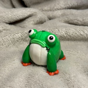 May include: A handcrafted, whimsical frog figurine. The frog is primarily green with a white belly and orange feet. It has large, expressive eyes and a slightly grumpy expression. The figurine is likely made of clay or a similar material.