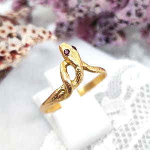 Puede incluir: Un anillo dorado con forma de serpiente, con ojos de piedras preciosas rojas. El cuerpo de la serpiente está entrelazado, creando un diseño único. El anillo se exhibe sobre una superficie blanca, con flores moradas borrosas en el fondo.