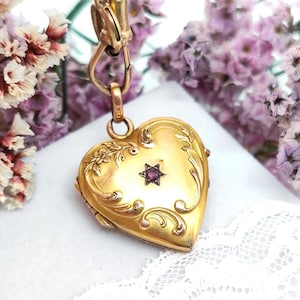 Könnte beinhalten: Ein goldfarbenes herzförmiges Medaillon mit aufwendigen floralen und scrollartigen Details. Ein kleiner, sternförmiger, lila Edelstein ist in der Mitte eingesetzt. Das Medaillon hängt an einer goldenen Kette und einem Verschluss. Der Hintergrund zeigt lila Blumen und weiße Spitze.