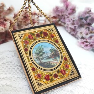 May include: A rectangular pendant with a miniature landscape painting. The pendant has a gold-colored frame with a black border and is adorned with red roses and small, sparkling accents. A gold chain suspends the pendant.