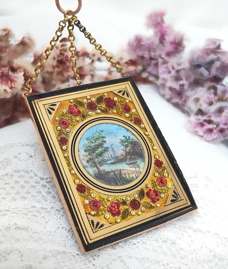 May include: A rectangular pendant with a miniature landscape painting. The pendant has a gold-colored frame with a black border and is adorned with red roses and small, sparkling accents. A gold chain suspends the pendant.