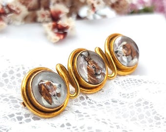May include: A gold-toned brooch featuring three circular settings, each with a miniature painting of a dog or rabbit. The paintings are detailed with brown and white fur. The brooch has a curved, swirled design connecting the settings.