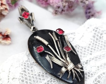 Peut inclure: Un pendentif vintage avec une base en émail noir et des détails en métal argenté. Le pendentif présente un motif floral avec des accents de pierres précieuses rouges. Le design comprend un bouquet de fleurs stylisées et des rubans. Le pendentif est posé sur un fond en dentelle.