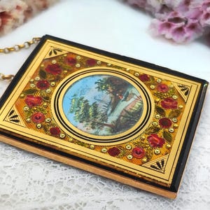 May include: A rectangular pendant with a gold frame and a black border. The pendant features a central oval image of a person in a landscape, surrounded by red roses and gold accents. A gold chain is attached to the pendant.