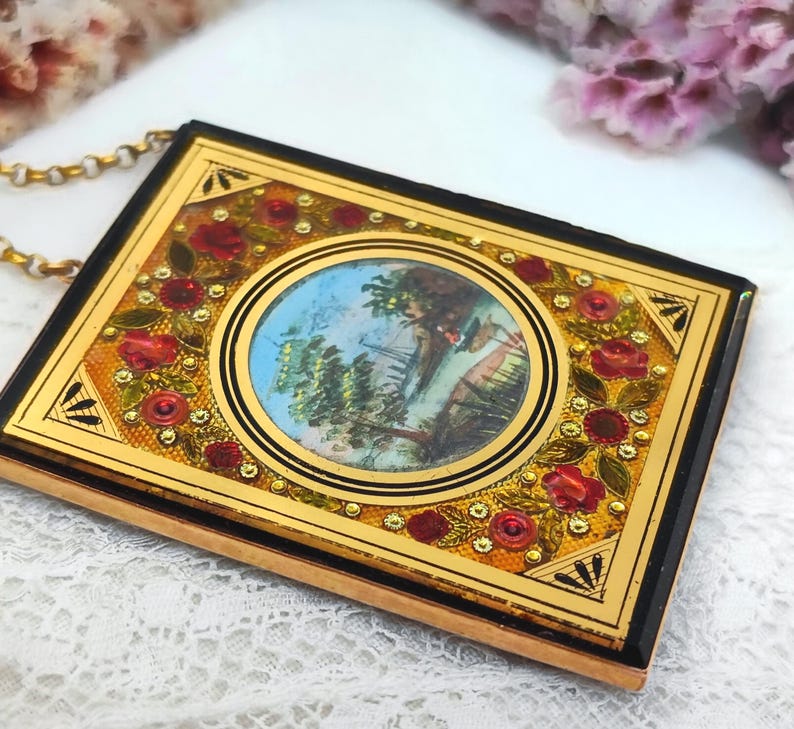 May include: A rectangular pendant with a gold frame and a black border. The pendant features a central oval image of a person in a landscape, surrounded by red roses and gold accents. A gold chain is attached to the pendant.