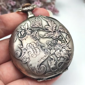 May include: A round silver-toned pocket watch featuring an intricate embossed floral design and a central shield-shaped cartouche with an engraved script letter B. The watch has a textured metallic winding crown at the top and a polished finish.