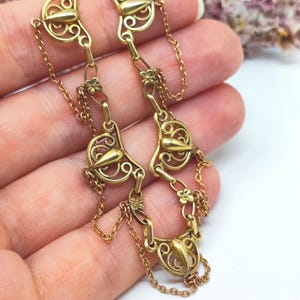 Op de afbeelding: Een goudkleurige metalen ketting met een reeks herhalende filigraan schakels. Elke schakel bestaat uit een centraal druppelvormig element omringd door ingewikkeld draadwerk, verbonden door kleine bloemmotieven met vier blaadjes en fijne kettinkjes.