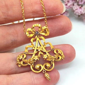 May include: A gold-colored pendant necklace with a bow and floral design. The pendant features a central purple gemstone and intricate detailing. The necklace is held by a person's fingers.