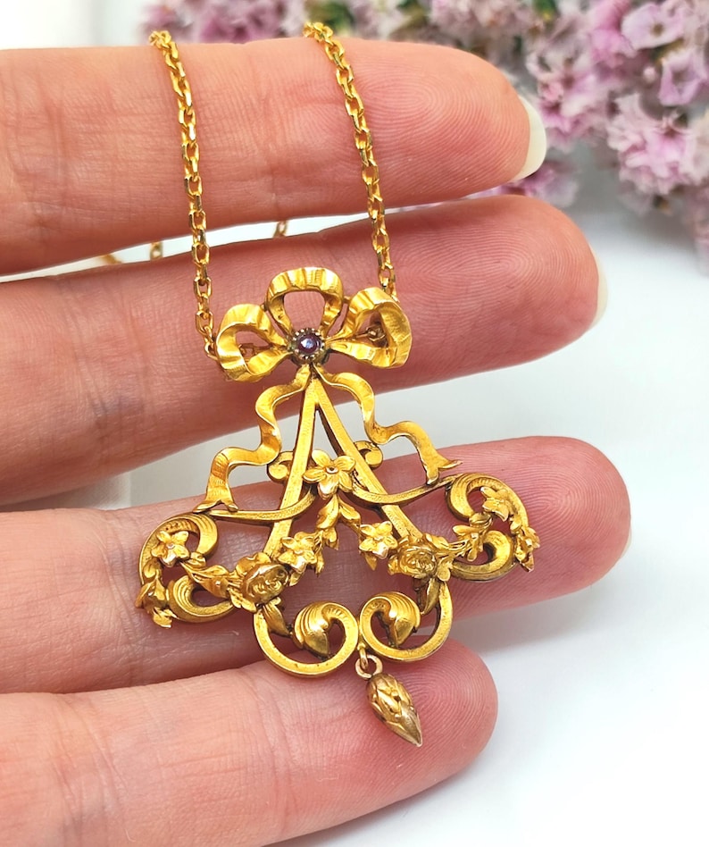 May include: A gold-colored pendant necklace with a bow and floral design. The pendant features a central purple gemstone and intricate detailing. The necklace is held by a person's fingers.