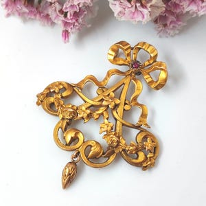 May include: A vintage gold-toned brooch with an ornate design. The brooch features a large bow at the top, a floral arrangement, and a dangling teardrop-shaped element. A small, round, red gemstone is set in the center of the bow. The brooch is set against a white background.