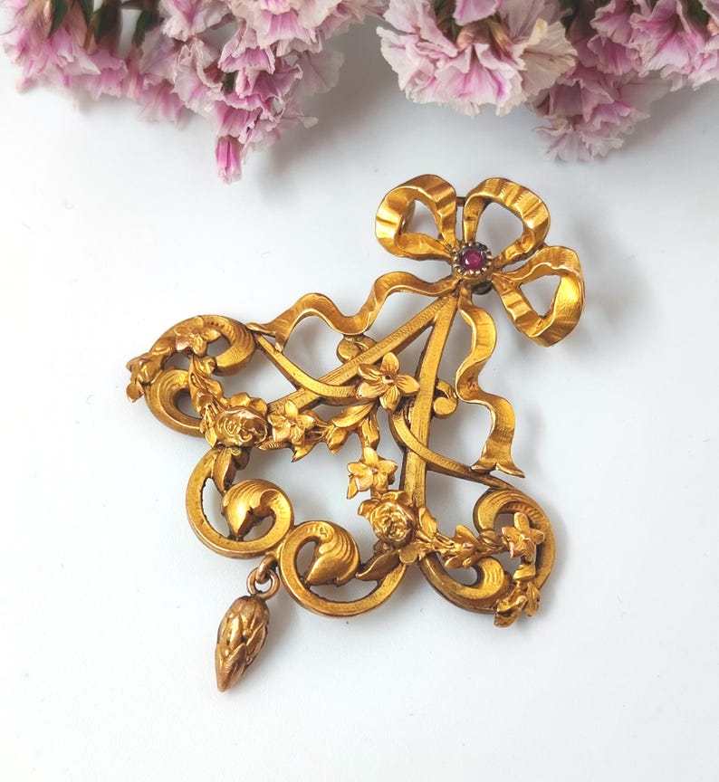 May include: A vintage gold-toned brooch with an ornate design. The brooch features a large bow at the top, a floral arrangement, and a dangling teardrop-shaped element. A small, round, red gemstone is set in the center of the bow. The brooch is set against a white background.