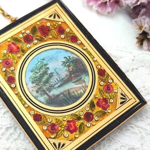 May include: A vintage rectangular compact with a gold frame and black edges. The compact features a central circular painting of a landscape scene. Red roses and rhinestones decorate the frame. A gold chain is attached.