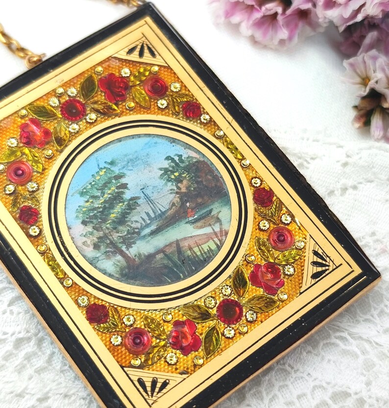 May include: A vintage rectangular compact with a gold frame and black edges. The compact features a central circular painting of a landscape scene. Red roses and rhinestones decorate the frame. A gold chain is attached.