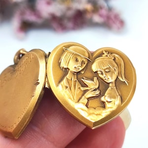 May include: A gold-toned heart-shaped locket featuring an embossed relief of two figures holding a bird. The left side of the open locket displays engraved cursive text. The metal surface has a matte finish with detailed raised imagery.