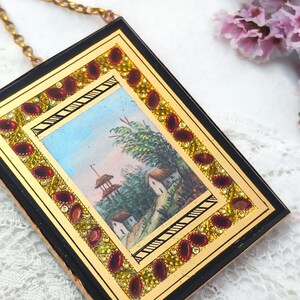 May include: A small, rectangular pendant with a miniature landscape painting. The painting is framed with gold and black borders, and adorned with red and yellow gemstones. A delicate gold chain is attached to the pendant.